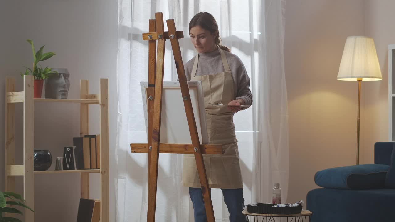 年轻女子的艺术爱好，女士正在客厅里用颜料作画，用画笔和画架作画布视频下载