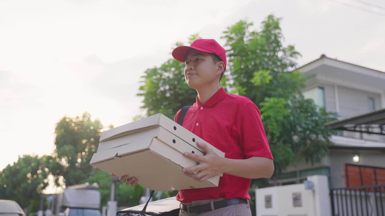 亚洲送货男工人穿着红色制服搬运食物，披萨给年轻漂亮的女顾客在房子前面。邮差和快递杂货配送服务理念视频素材