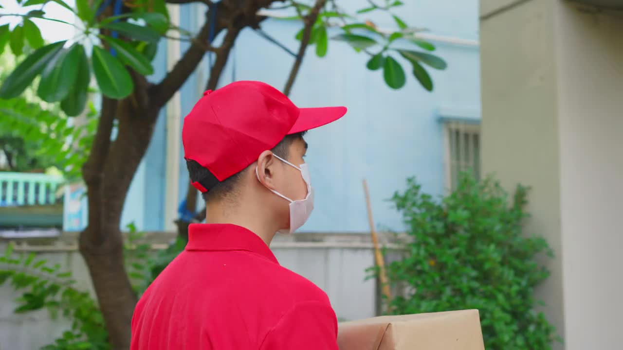 亚洲的邮递员、快递员戴着口罩把小箱子送到顾客家门口。戴口罩的人预防covid - 19、corana病毒情感爆发。送货上门的购物理念。视频下载