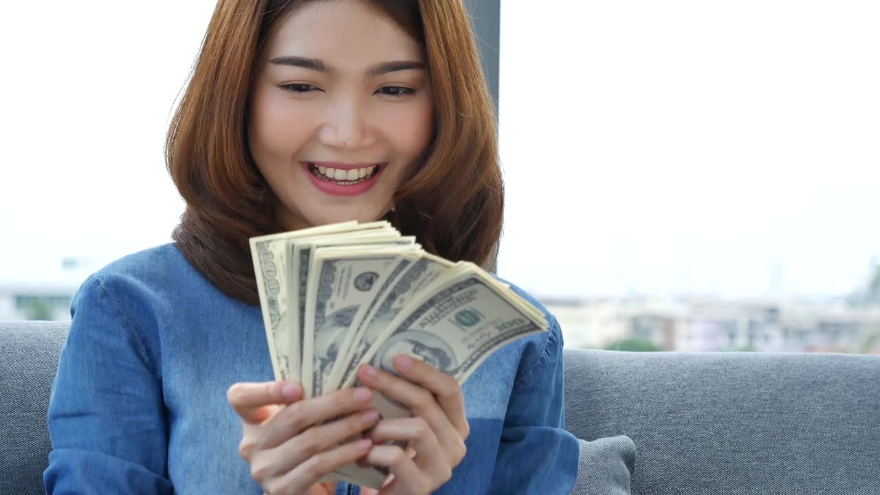 快乐的年轻亚洲妇女拿着美元的钱，扔在空中庆祝幸福舞蹈财富彩票钱雨点。赢家成功的商业女人扔现金流快乐的钱笑脸视频素材