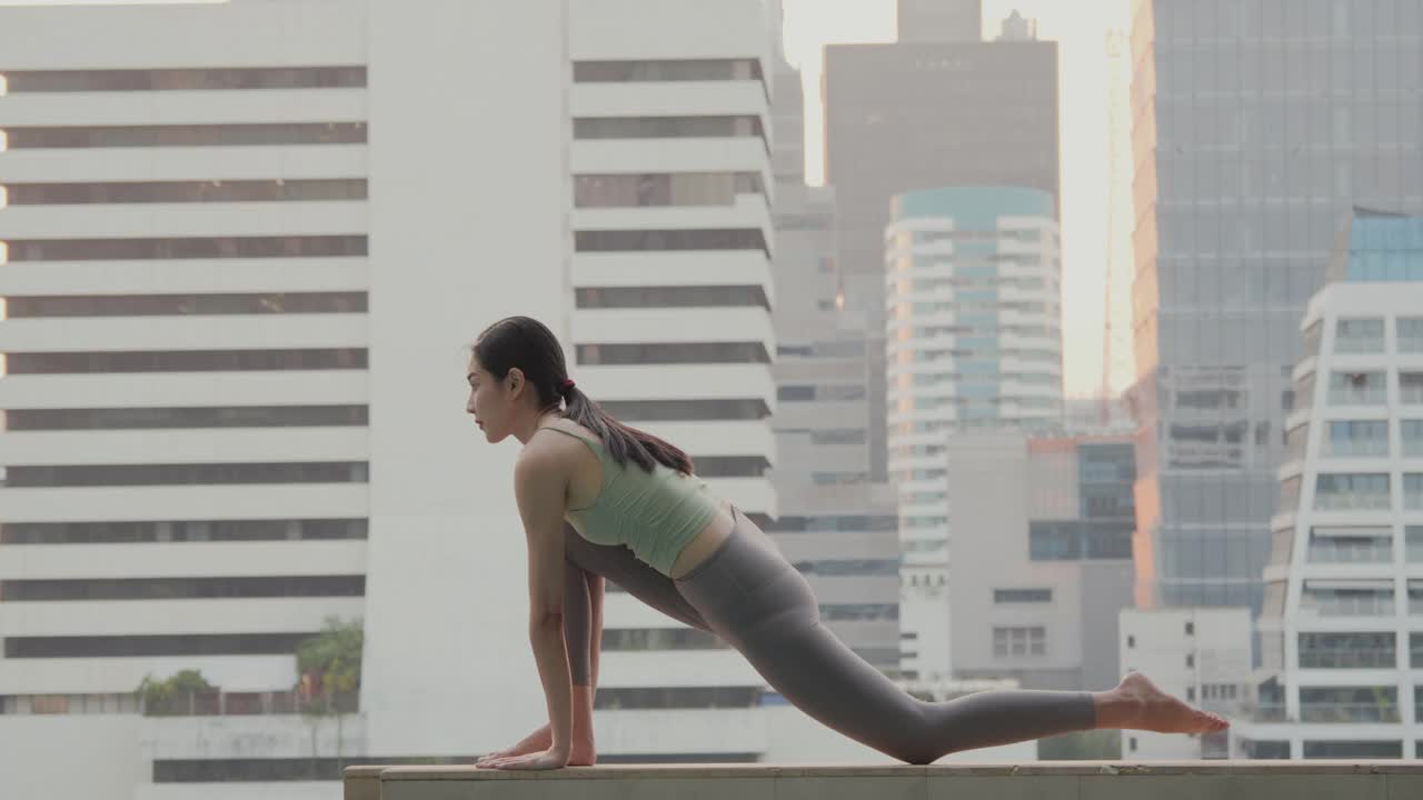 一个年轻漂亮的女人长头发做瑜伽练习和瑜伽姿势叫战士姿势在摩天大楼的露台上感觉放松和和平。视频下载
