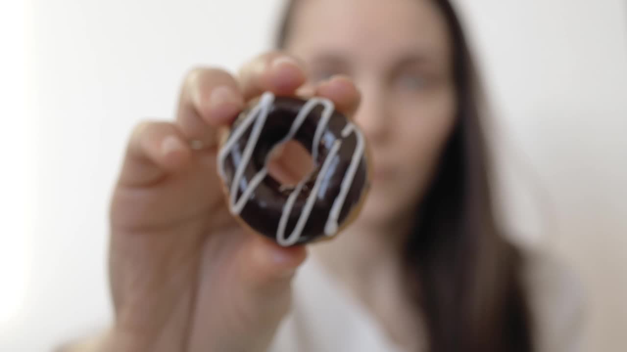 一个有着深色直发和蓝眼睛的女人手里拿着一个涂满黑巧克力和白色糖霜的甜甜圈。特写镜头。饮食概念视频素材