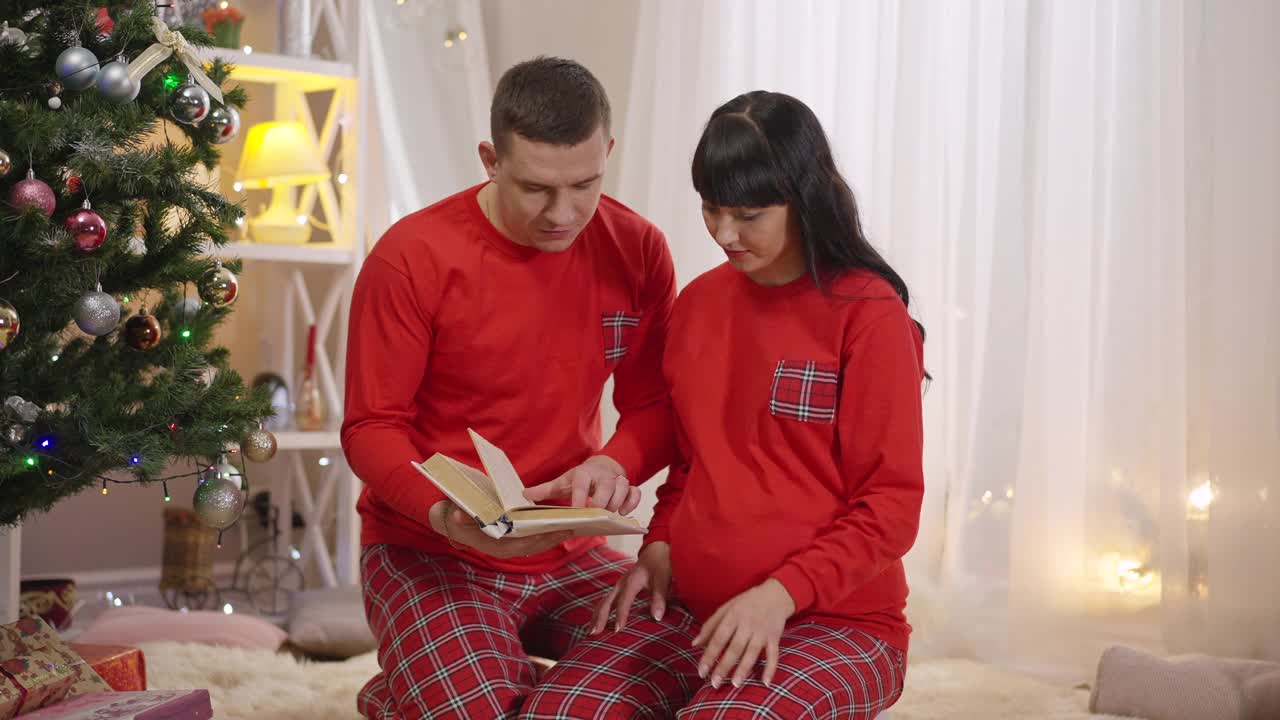 一对快乐的夫妇穿着相似的红色睡衣坐在家里的圣诞树旁看书。积极怀孕的白人妇女和微笑的男子享受休闲新年。团结的快乐视频素材