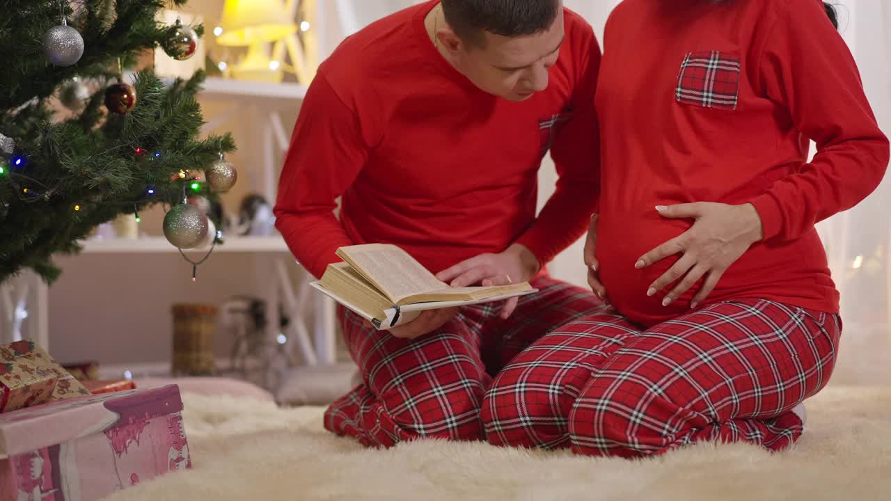 爱丈夫对着肚子看书，亲着胎儿。幸福的白人男子在室内享受圣诞节家庭晚会的肖像。新年与怀孕视频素材