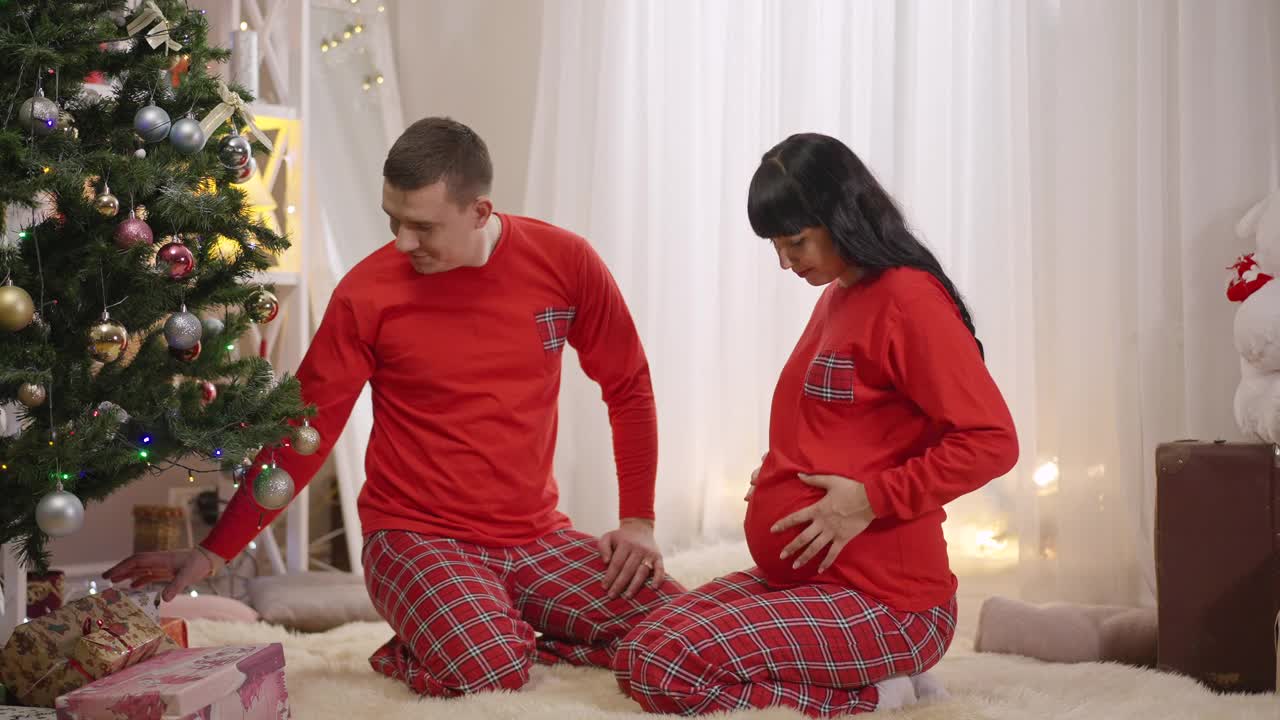 微笑的丈夫给兴奋的怀孕的妻子在红色睡衣圣诞礼物。幸福相爱的白人男人和期待的女人在家庆祝除夕。慢动作视频素材