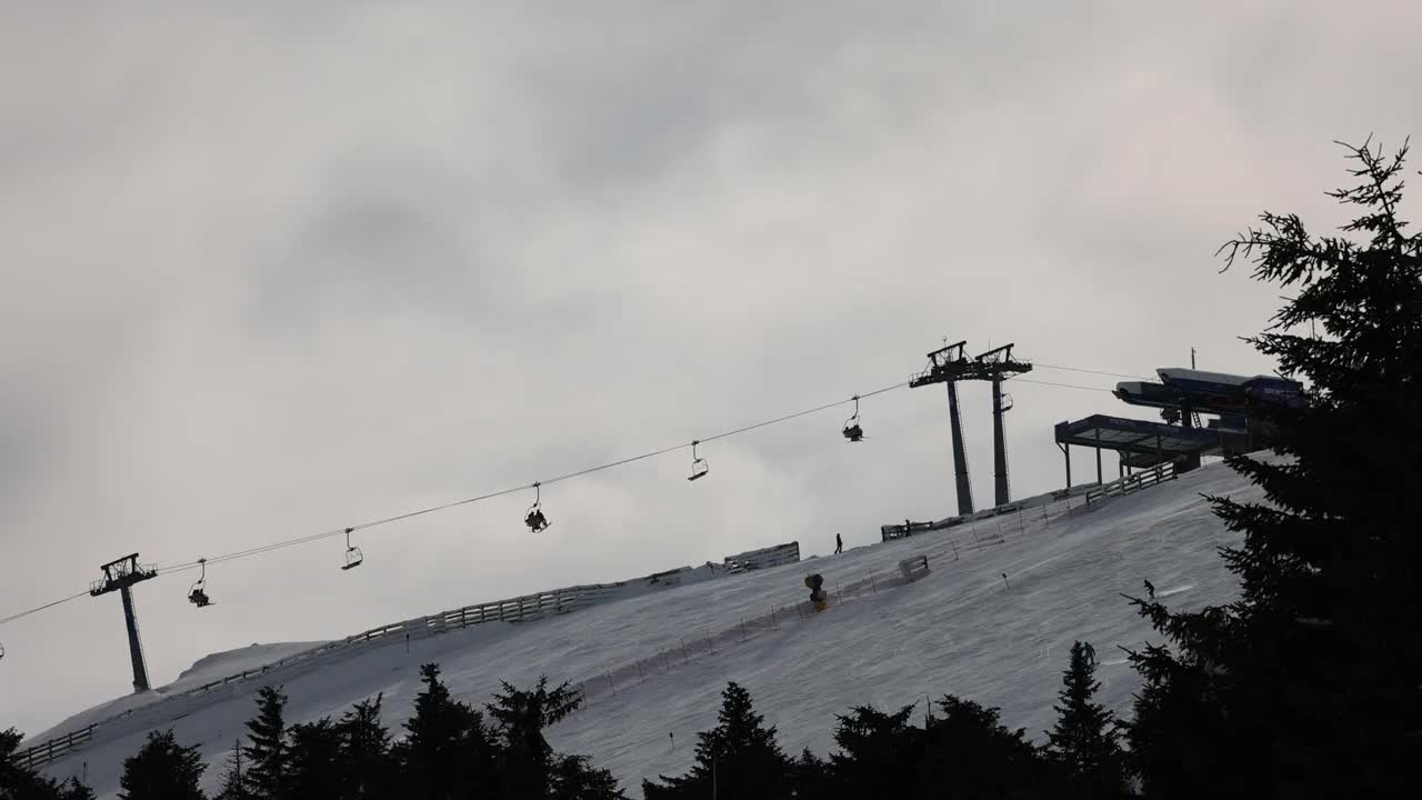 在阴天的冬季，在索道上移动的升降机和在滑雪道上滑雪的滑雪者视频下载