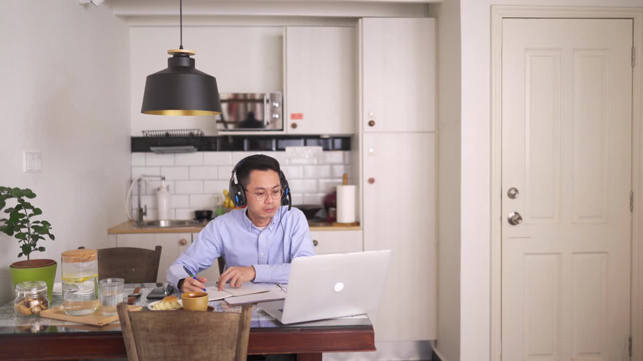 亚洲华人中年男子面带微笑，戴着耳机在用餐区使用笔记本电脑参加在线会议视频素材