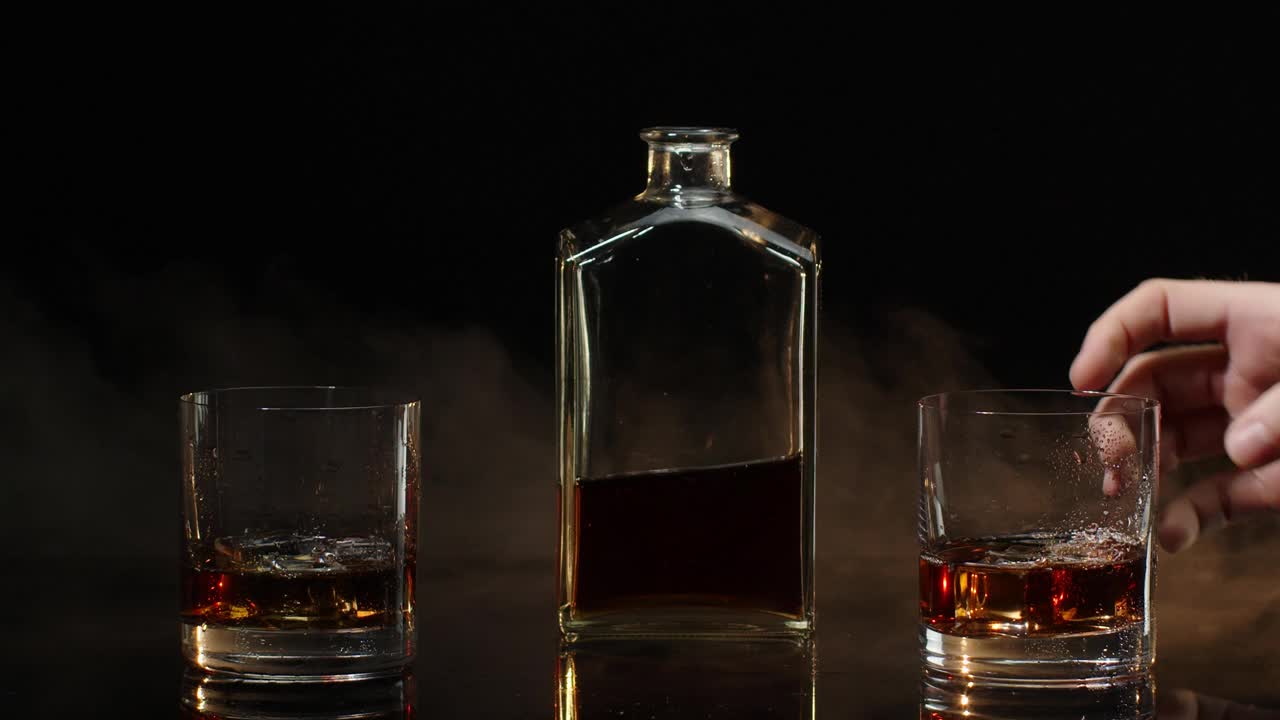 双手与干邑威士忌杯与冰块使干杯祝酒词在黑色背景视频下载