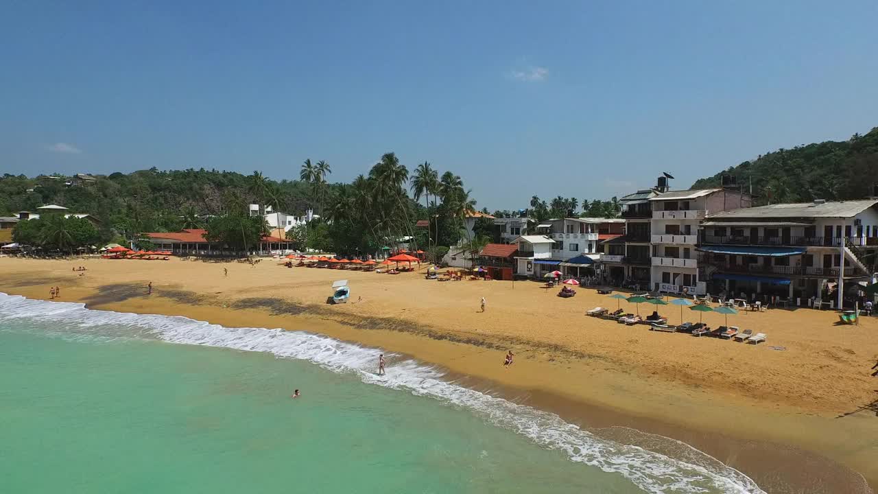 在一个阳光明媚的日子里，有人们，船，海岸建筑，树木，温柔的海浪，和明亮的蓝天——乌纳瓦图纳，斯里兰卡视频素材