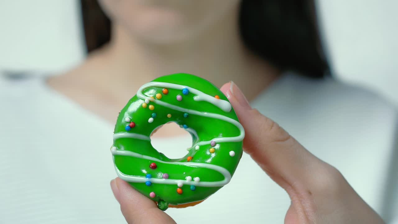 一个披头散发、身穿白色衣服的女人，手里拿着一个有绿色水果糖霜和多种颜色糖霜的甜甜圈。特写镜头。饮食概念视频素材