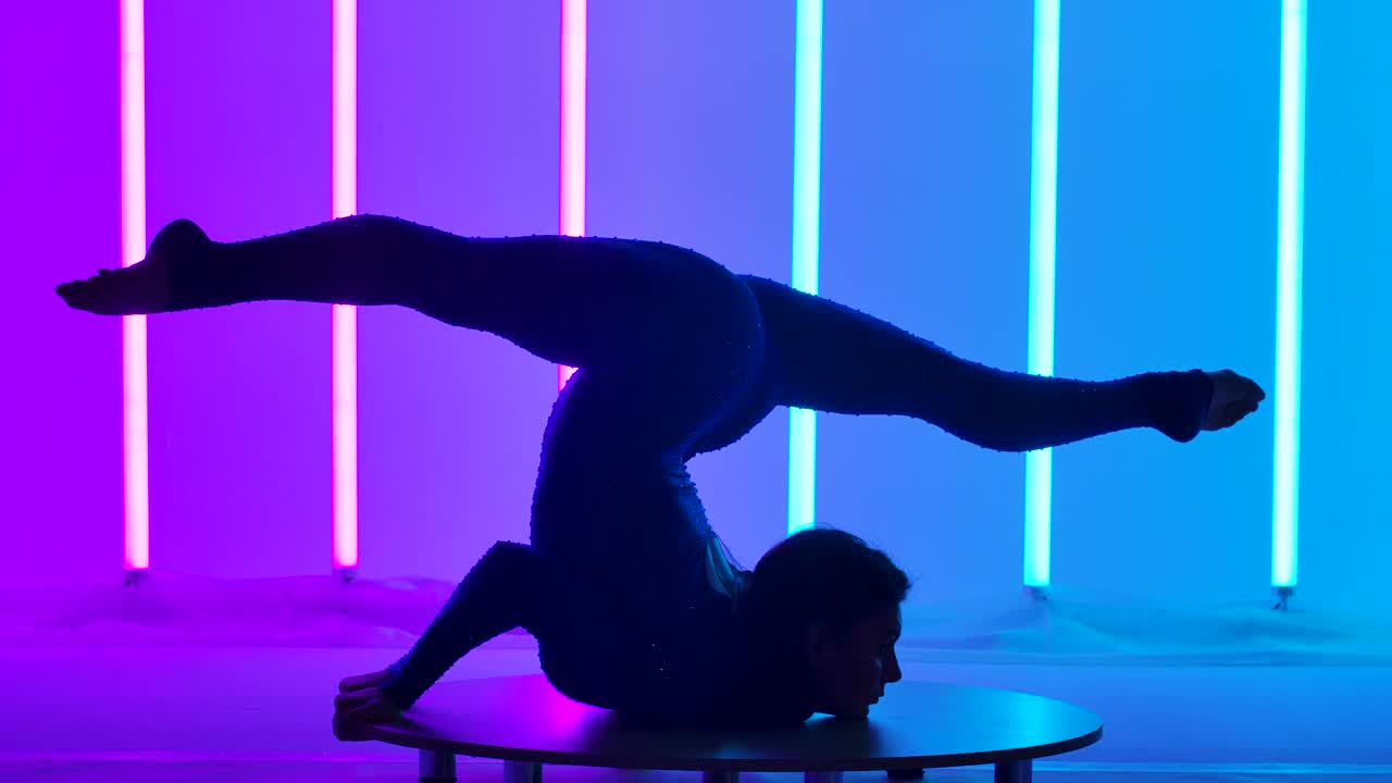 在粉色和蓝色霓虹灯的背景下，体操运动员在演播室里。女孩做了一个胸部站立，伸直双腿，坐在麻绳上。轮廓。缓慢的运动。近距离视频下载