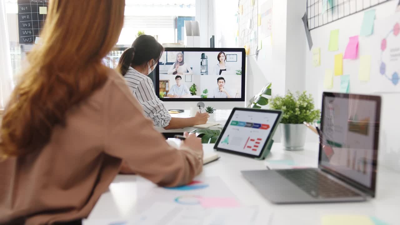 在新常态的办公室里，亚洲商务人士戴着口罩在桌面上与同事们讨论视频通话会议的方案。视频素材