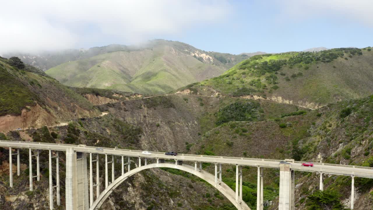 从横跨两座山的1号公路大桥向太平洋的空中倒车，背景中有云、蓝天、汽车和起伏的山丘——大苏尔，加利福尼亚视频素材