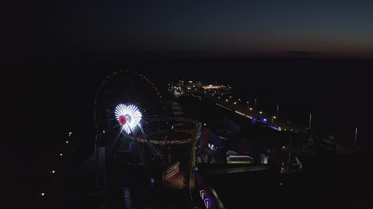 航拍拍摄心形的摩天轮在码头照明，无人机在夜间飞过著名的圣塔莫尼卡码头视频下载
