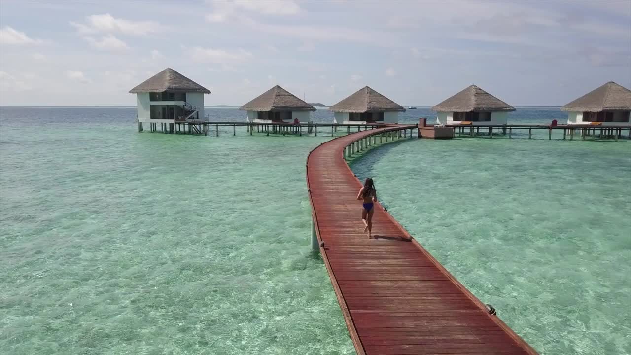 航拍的女人在码头上跑步的水平房，无人机上升向前在豪华度假村的绿松石海对天空-瓦都，马尔代夫视频下载