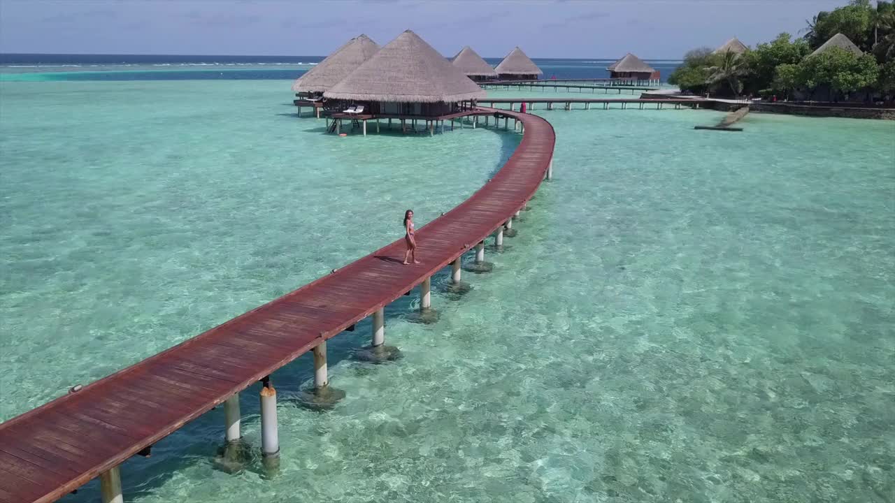 航拍的女人在水上小屋的码头上奔跑，无人机向前飞行在豪华度假村的绿松石海对天空-瓦都，马尔代夫视频下载