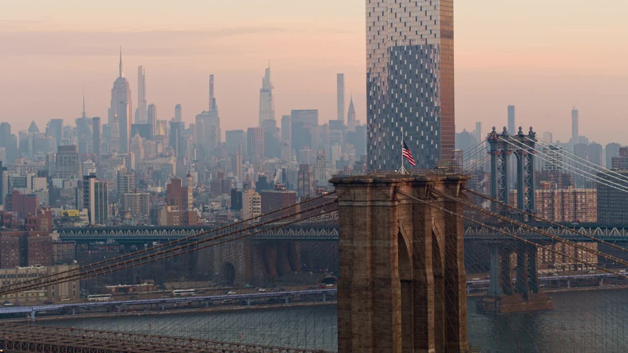 近距离鸟瞰布鲁克林大桥塔顶飘扬的美国国旗，以曼哈顿天际线为背景，清晨时分。无人机视频与向后相机运动。视频素材