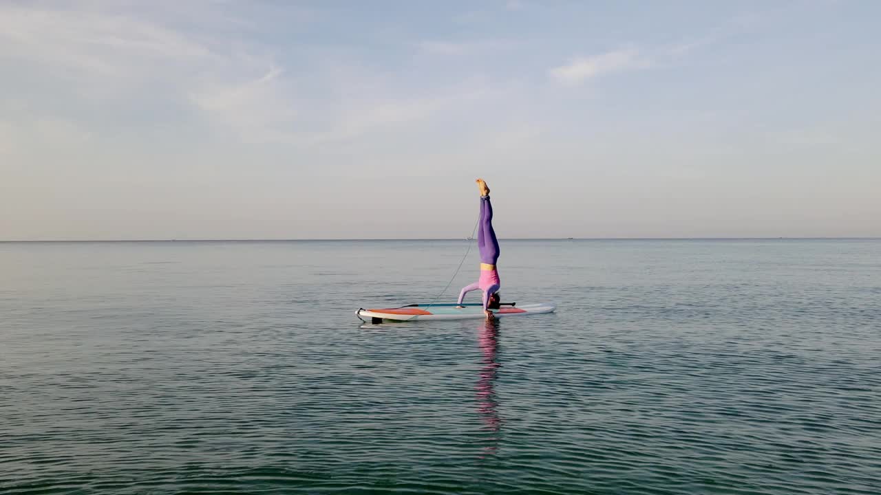 无人机航拍，锻炼女性早上在海滩户外伸展身体。早晨在冲浪板上练习瑜伽，海面上有柔和的阳光。视频下载