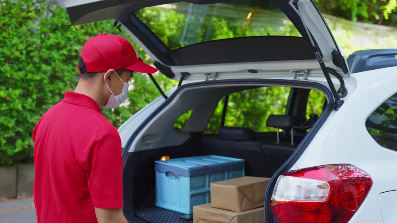 亚洲快递员戴着口罩穿着红色制服，搬运包裹盒子给女顾客走到房前。新冠肺炎期间的邮差及快递杂货送货服务。视频素材