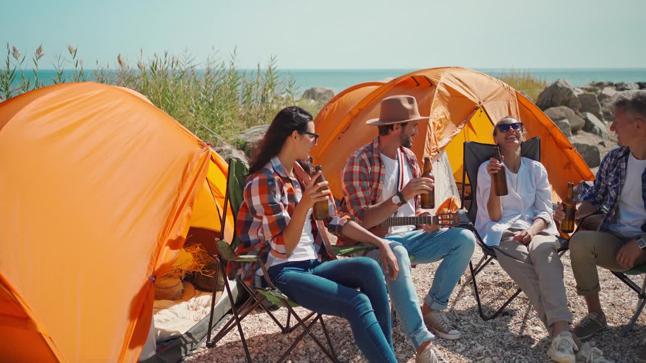 一群快乐的朋友一起喝啤酒，一起在海滩上玩。成年人在海边露营，喝啤酒，一起享受视频下载