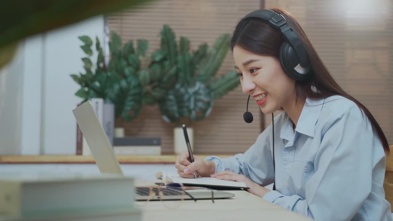 年轻美丽的亚洲女性戴着耳机使用笔记本电脑在家里办公视频下载