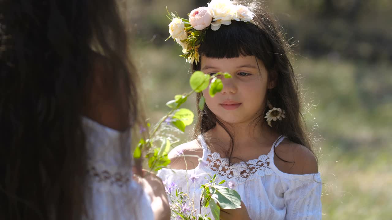 特写的小高加索学前班女孩与长棕色头发和蓝色眼睛在花圈看着她的姐姐收集花束在自然背景。4k 50fps慢动作视频下载