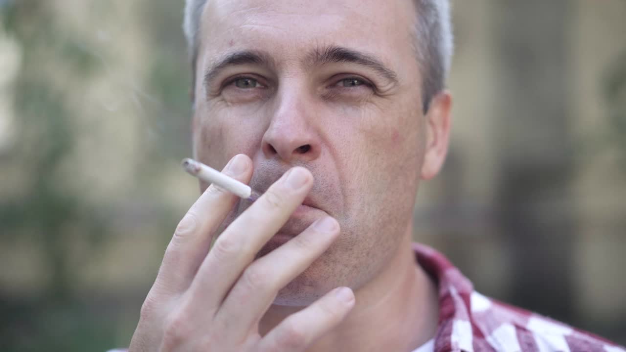 一个自信的灰白头发的白人男子在户外抽烟，一边看着镜头一边吹着烟。特写的人有坏习惯上瘾。烟草成瘾视频素材