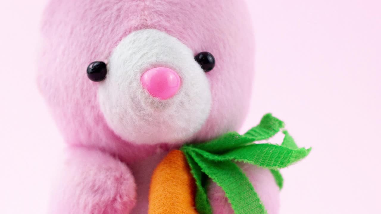粉色复活节毛绒兔子玩具和一个橙色的小胡萝卜坐在一个柔和的背景，特写，慢动作。概念为宗教节日春天。视频下载