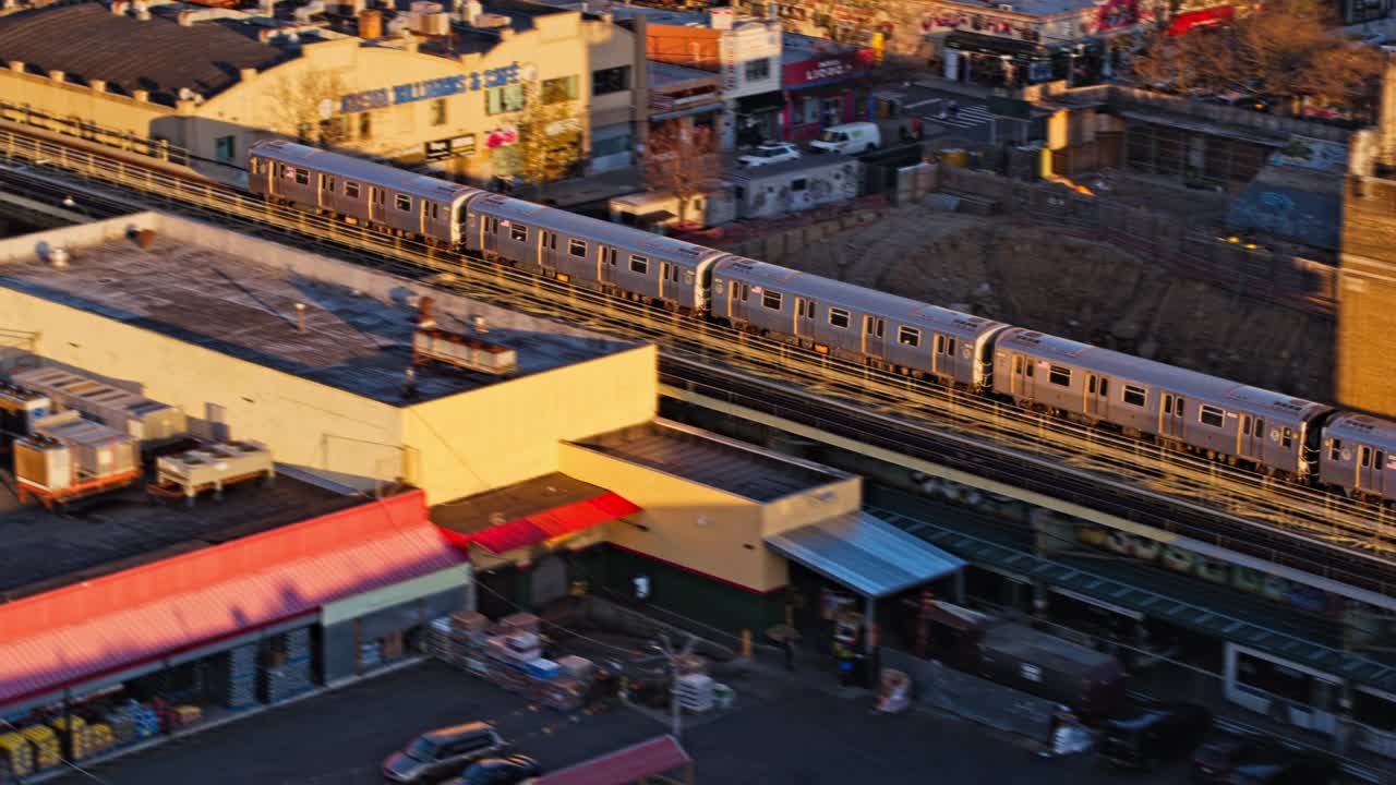 在一个阳光明媚的日子里，火车在纽约布鲁克林默特尔大道的高架地铁线上经过。无人机视频倾斜移位效果，与前摇相机运动，跟踪列车。视频下载