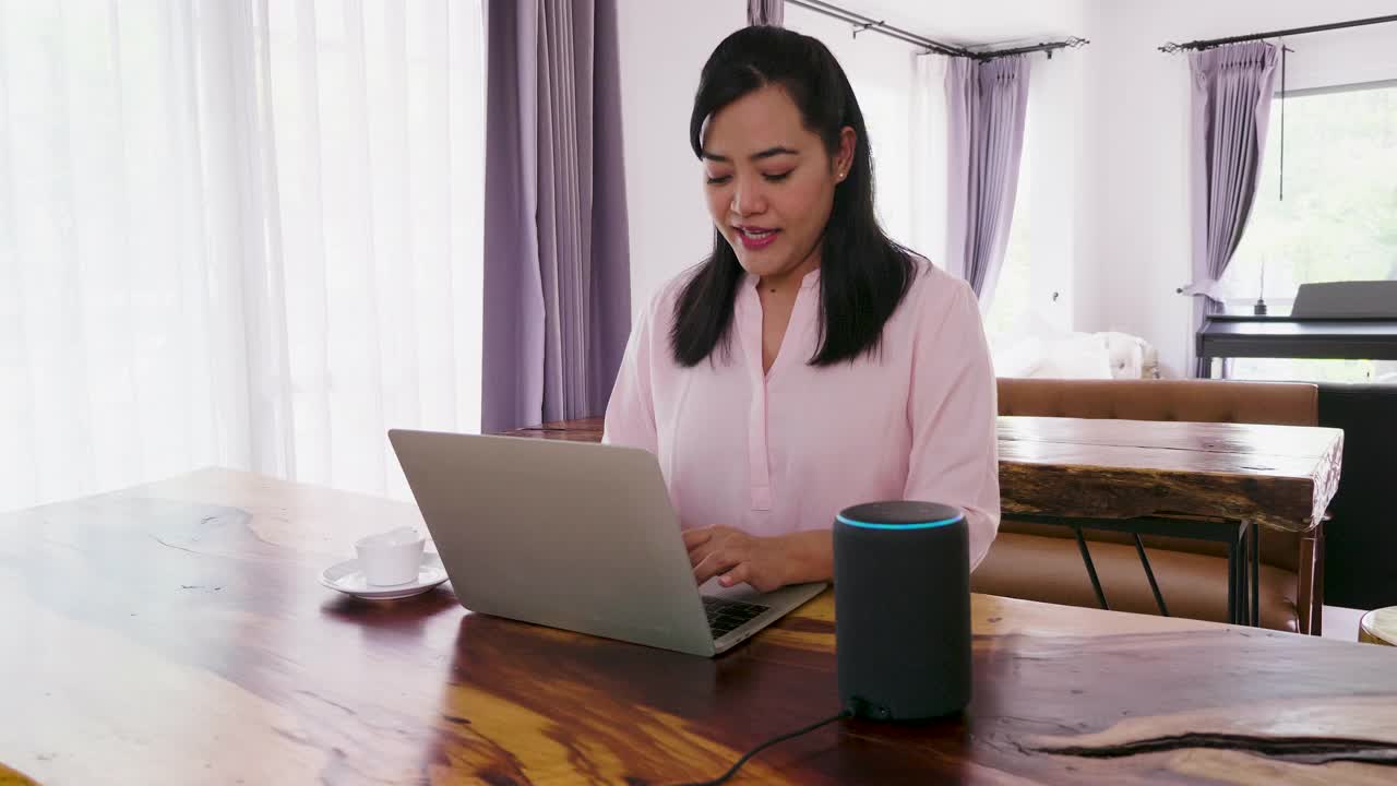 亚洲成熟女性在家里的客厅里使用笔记本电脑和智能扬声器工作视频素材
