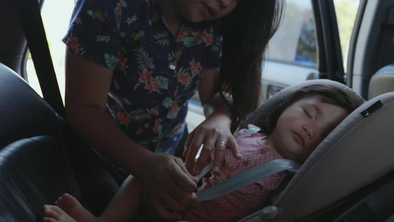 亚洲母亲扣安全带给她的女儿，当她睡在汽车座位上的公路旅行。视频下载