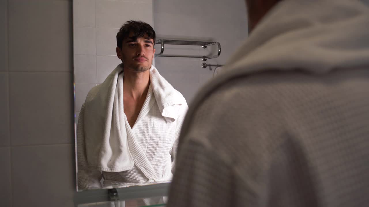 一个穿着白袍的年轻人站在浴室里靠近镜子的地方刷牙视频素材