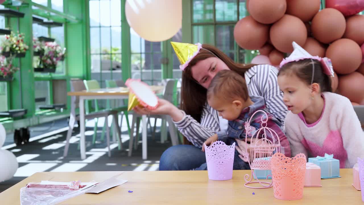 当她的小女儿在玩派对装饰时，一位拉丁妈妈在玩她1岁的女儿和一顶庆祝帽视频下载