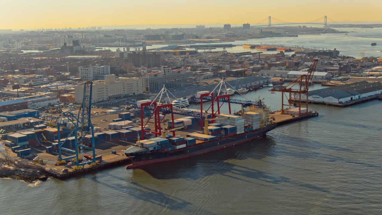 日落时分，一艘工业集装箱船正在纽约布鲁克林东河的商业码头装载货物。无人机制作的视频与电影全景轨道摄像机运动。视频下载