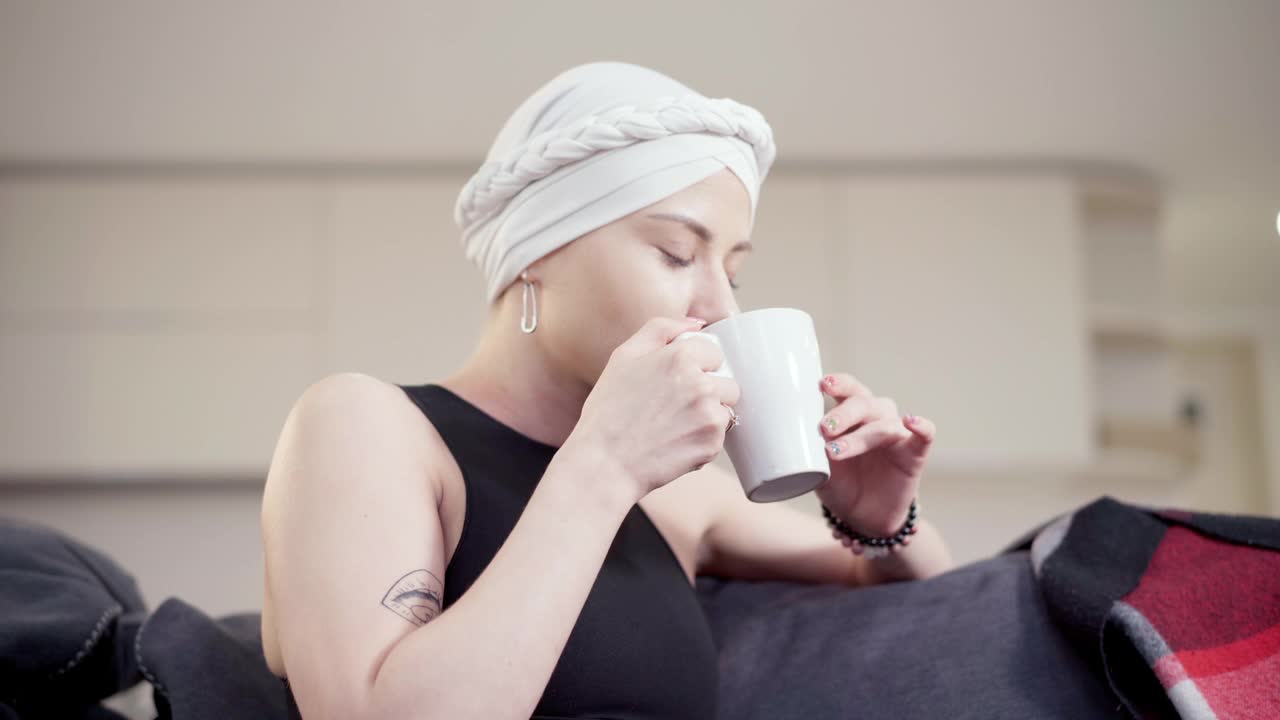 侧面的苗条的年轻女子喝着茶或咖啡有严重的头痛触摸头部。一个可怜的白人女士坐在家里的沙发上休息。疾病症状和保健。视频素材
