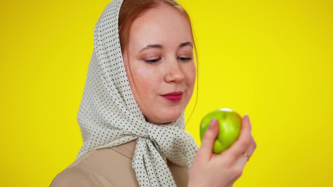 美丽的年轻红发女子绿色眼睛吃健康多汁的苹果在慢镜头。特写的苗条的白人女士享受健康的水果在黄色的背景视频素材