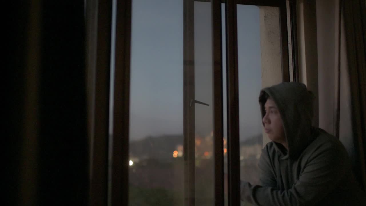 亚洲华人中年男子看窗外从他的家在日落与严肃的面部表情与帽衫视频素材