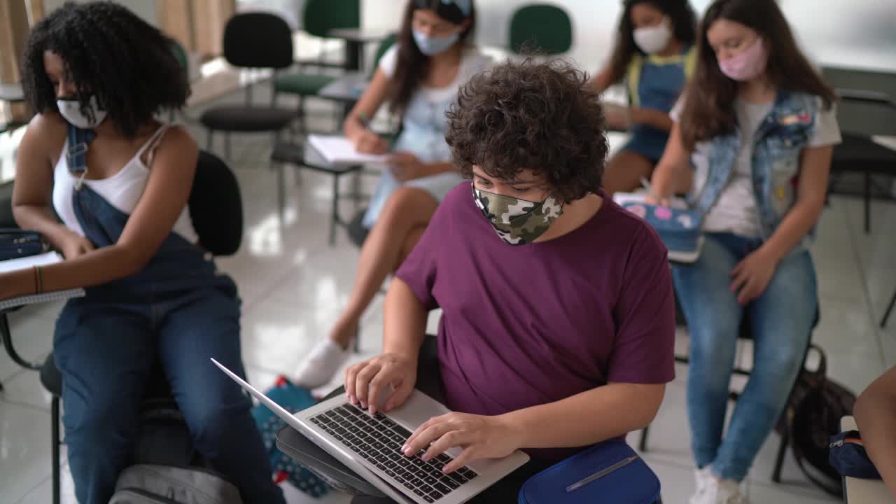 一个十几岁的男孩戴着口罩在教室里使用笔记本电脑的肖像视频下载