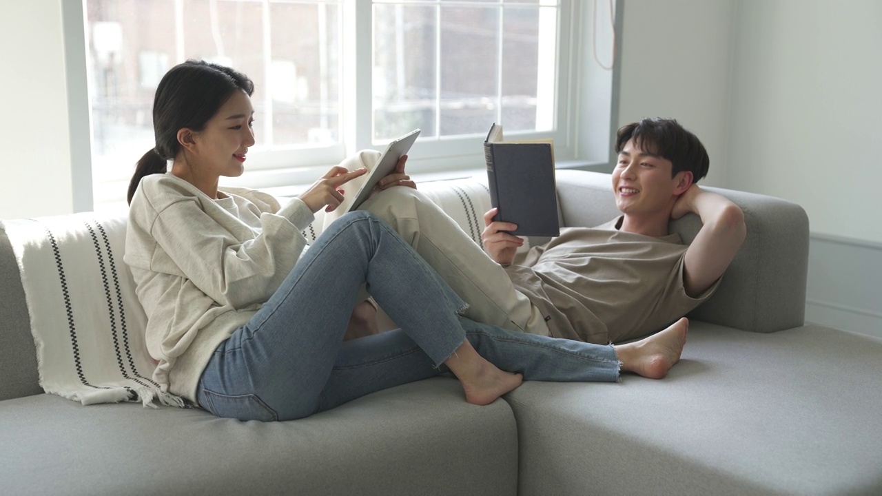 一对夫妇——年轻男子躺在沙发上看书，年轻女子靠在沙发上使用平板电脑视频下载