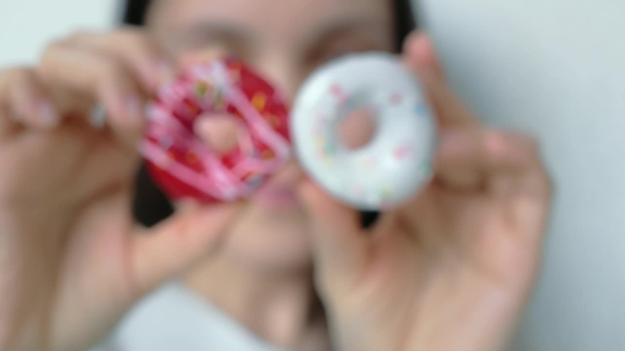 一个女人的双手展示了两个甜甜圈。一个上面覆盖着白色条纹的红色糖衣，另一个上面覆盖着白色糖衣，上面撒着多种颜色的糖屑。特写镜头。饮食概念视频素材