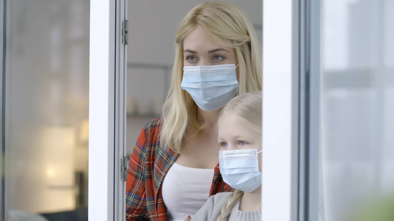 悲伤的母亲和小女儿戴着医用口罩望向窗外，是covid-19视频素材