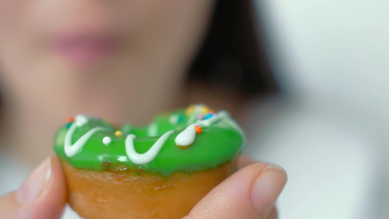 一个女人手里拿着一个油炸圈饼。甜甜圈上覆盖着绿色的糖霜、白巧克力和糖果屑。女孩吃了一口被咬掉的甜甜圈。特写镜头。饮食概念视频素材