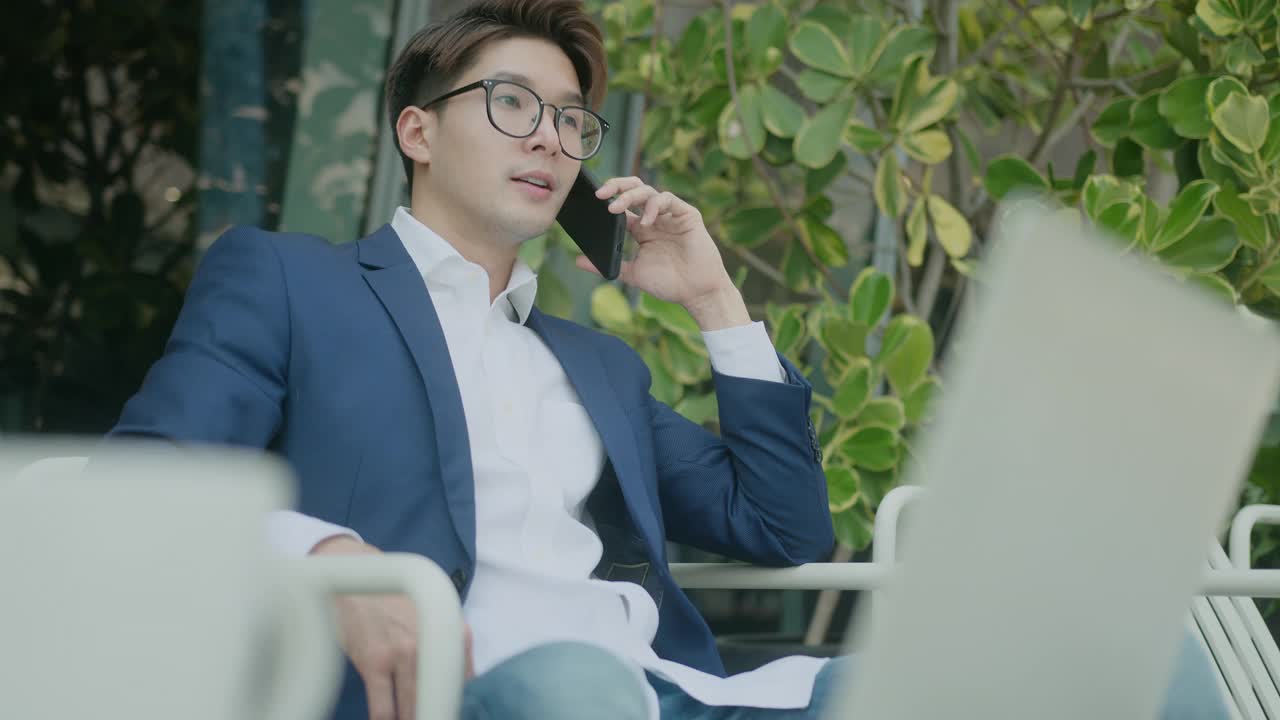 年轻聪明迷人的亚洲自信男性穿着正装，布，手持智能手机，在写字楼庭院的现代花园与客户沟通，随意咨询视频下载