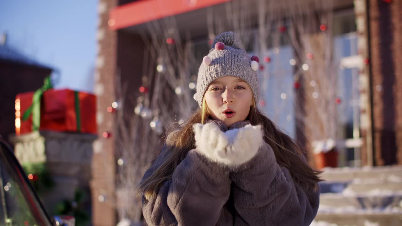 微笑的女孩在针织帽和皮草大衣吹展示从手上的圣诞装饰品。欢乐的少女吹蓬松的雪从手户外新年假期。视频素材