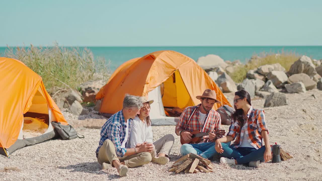 全身照:两对好朋友坐在帐篷附近的海滩上，谈笑风生，玩尤克里里琴视频下载