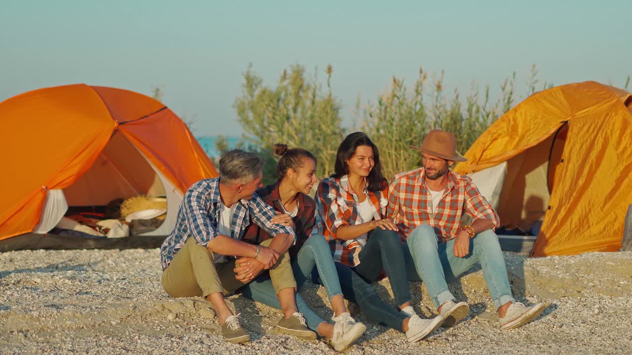 四个欢快迷人的朋友旅行者坐在海滩附近的橙色帐篷，说说笑笑视频下载