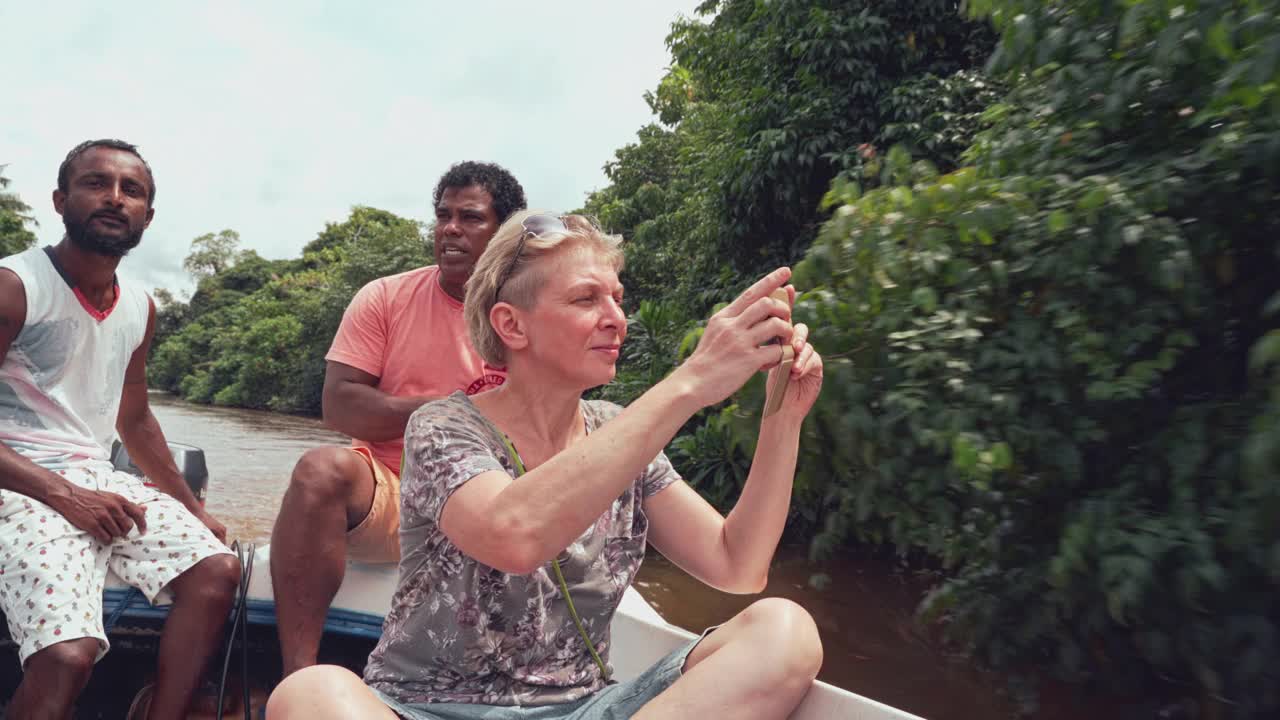 斯里兰卡本托塔河上，一名留着短发的白人老年妇女，在两名当地导游的陪同下，在一艘船上用她的智能手机拍照。视频下载