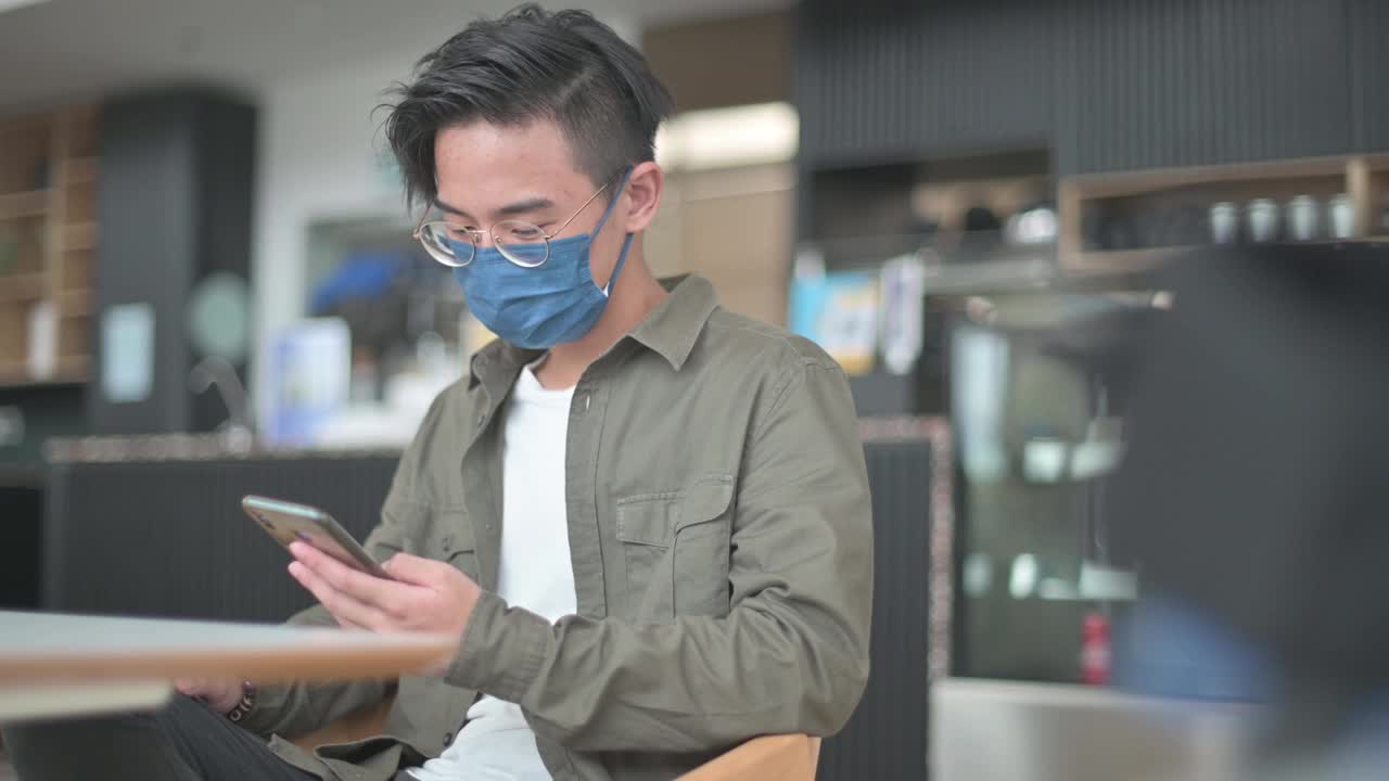 一名戴着保护面罩的亚裔中国男孩坐在咖啡馆里刷屏，查看社交媒体视频素材