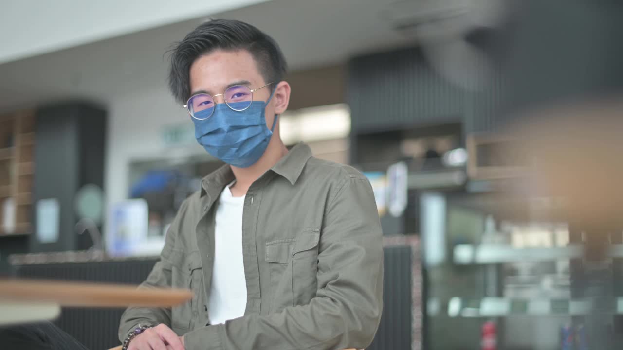 一名亚裔中国少年戴着防护面具坐在咖啡馆里看镜头视频素材