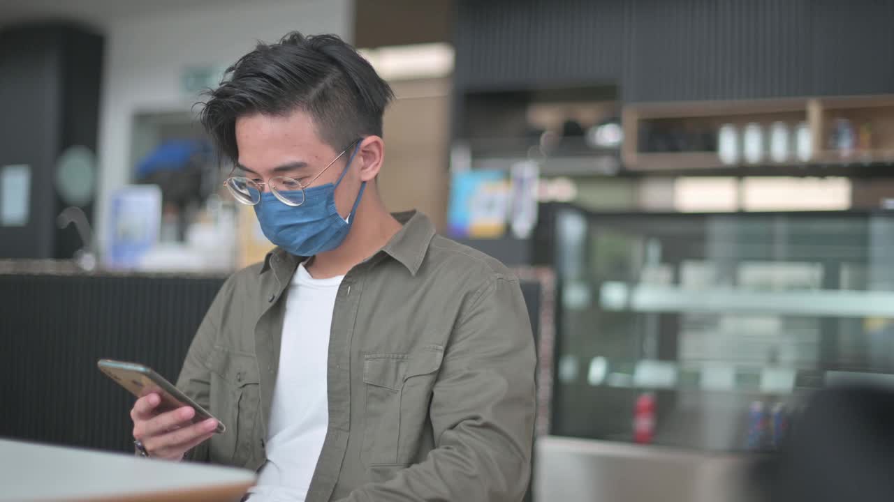 一名戴着保护面罩的亚裔中国男孩坐在咖啡馆里刷屏，查看社交媒体视频素材